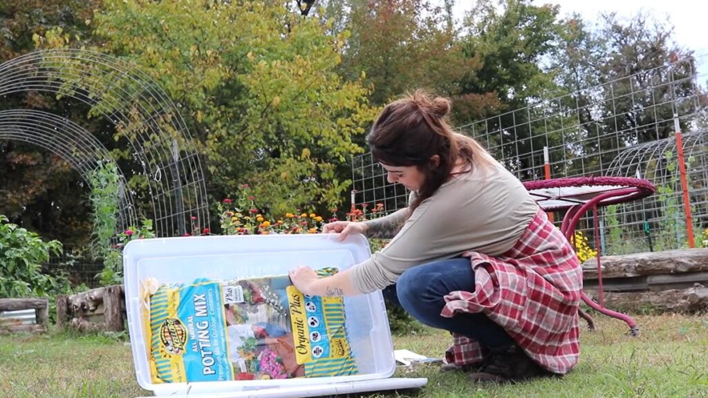 A woman crouched in the garden with a large plastic bin and a bag of soil.