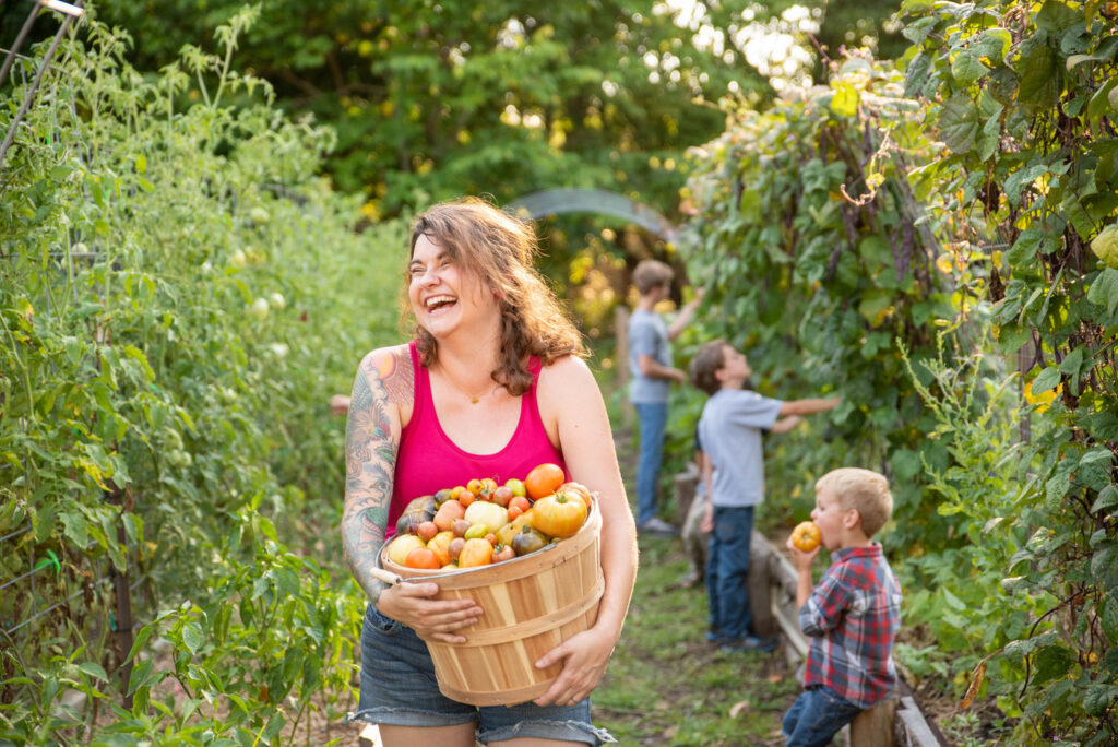 A woman carrying a large basket filled with fresh picked tomatoes. Her family is in the background.