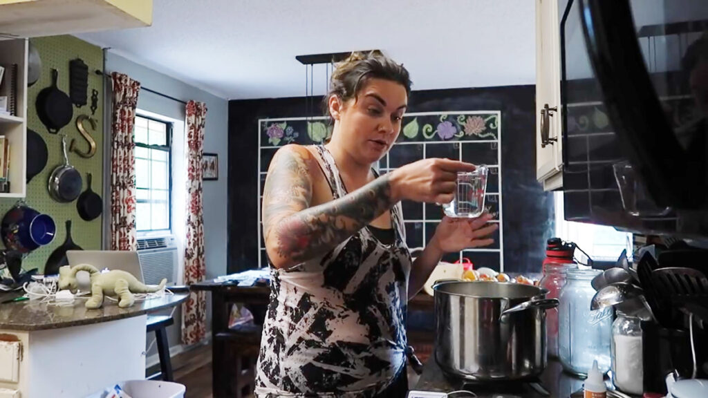 A woman adding a measuring cup of water with rennet into a pot of milk to make homemade mozzarella cheese.