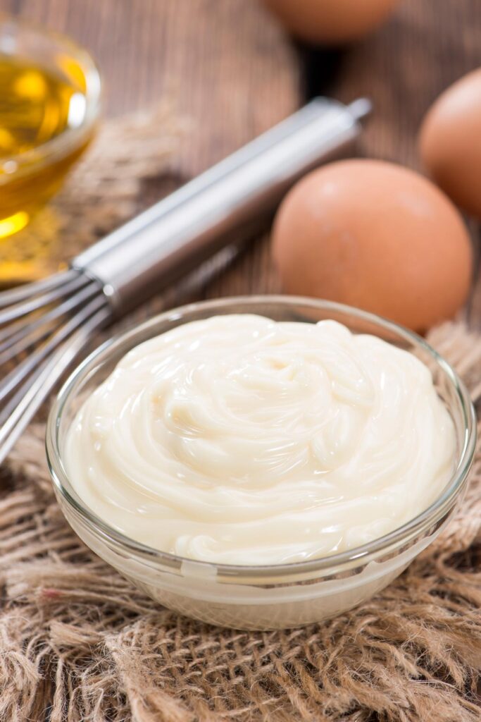 Homemade mayonnaise in a glass bowl with eggs and a whisk in the background.