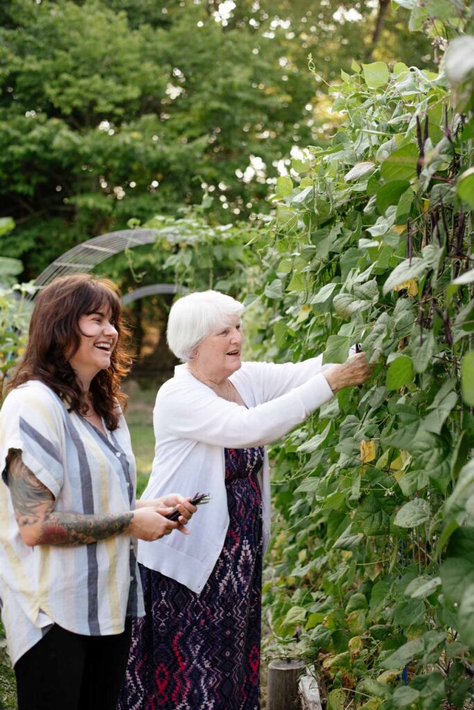 A younger and older woman picking beans off a climbing trellis.