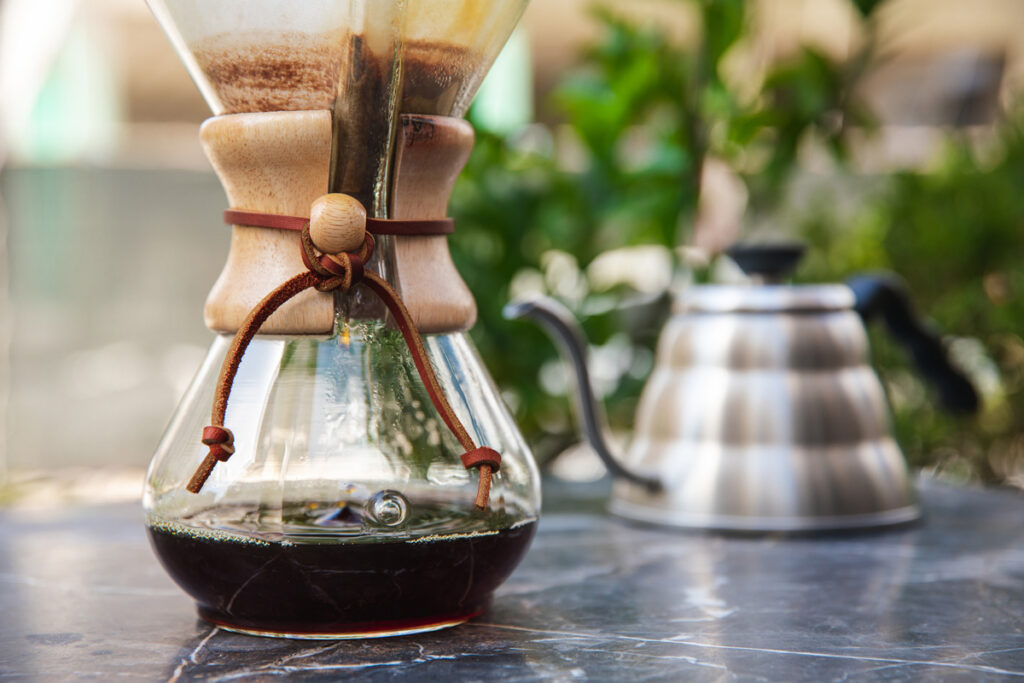 Close up of a Chemex coffee pot and a teapot of water in the background.