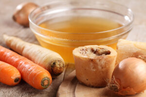 Bone broth in a glass bowl with bones, carrots and onions on a counter.