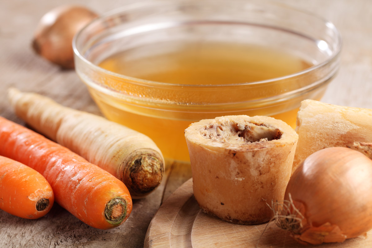 Bone broth in a glass bowl with bones, carrots and onions on a counter.