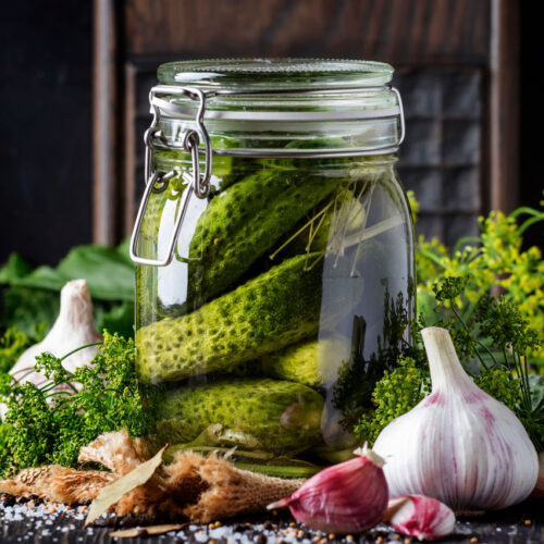 A jar of pickles with dill, garlic and other herbs around it.