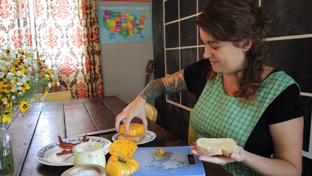 A woman adding a large slice of a yellow tomato to a sandwich.