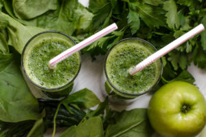 Two glasses of green juice with greens and an apple on the table.
