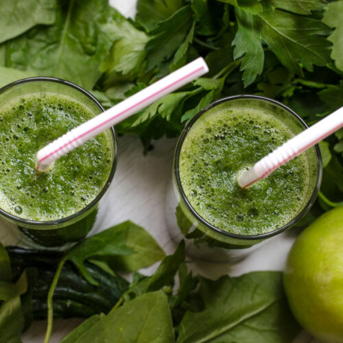 Two glasses of green juice with greens and an apple on the table.