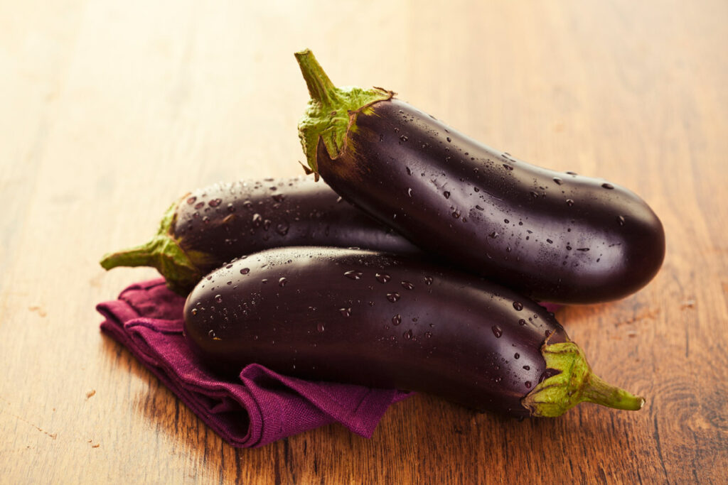 Three eggplant on a purple towel on a wooden counter.