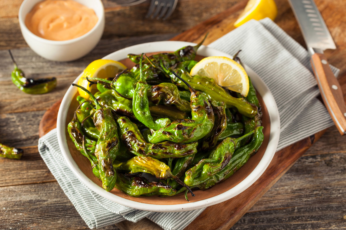 Blistered shishito peppers in a bowl with lemon wedges and a bowl of aioli.