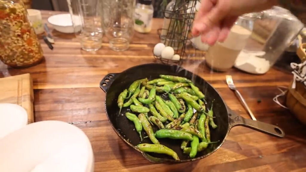A hand sprinkling salt over blistered shishito peppers in a cast iron pan.