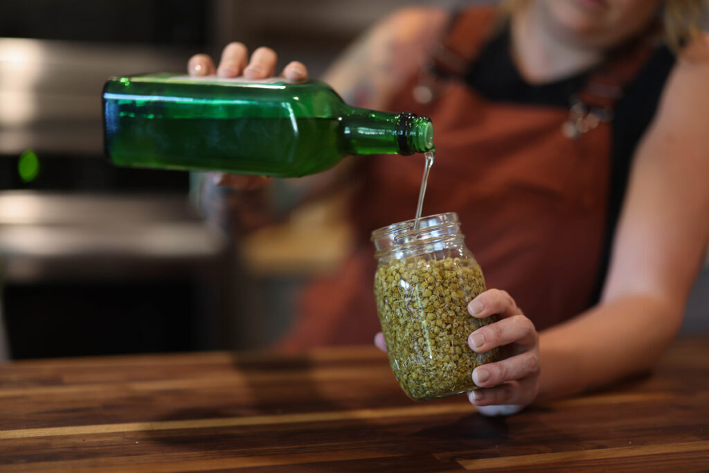 Dried herbs in a Mason jar with a woman pouring oil over the top.