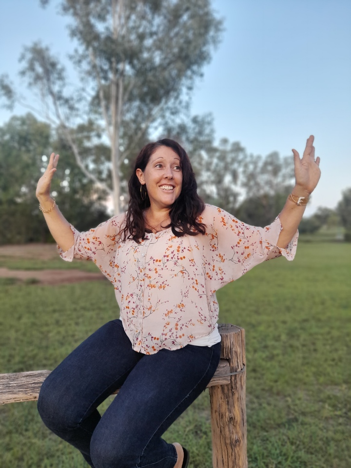 A woman sitting on a wooden fence with her arms raised.